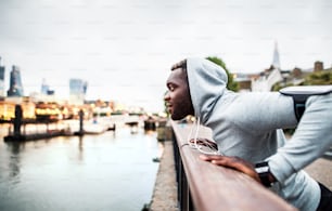 Junger sportlicher schwarzer Mann Läufer mit Smartwatch, Kopfhörern und Smartphone in einem Armband auf der Brücke in einer Stadt, ruhend.