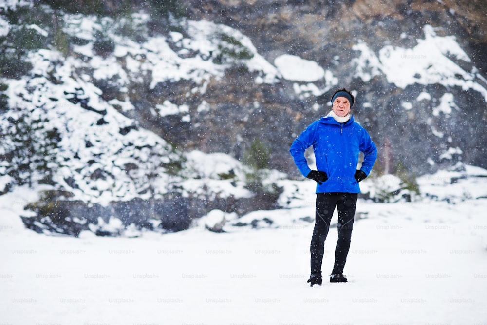 A full-length portrait of senior man standing after the run in winter nature, hands on hips. Copy space.