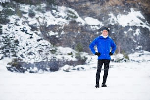 A full-length portrait of senior man standing after the run in winter nature, hands on hips. Copy space.