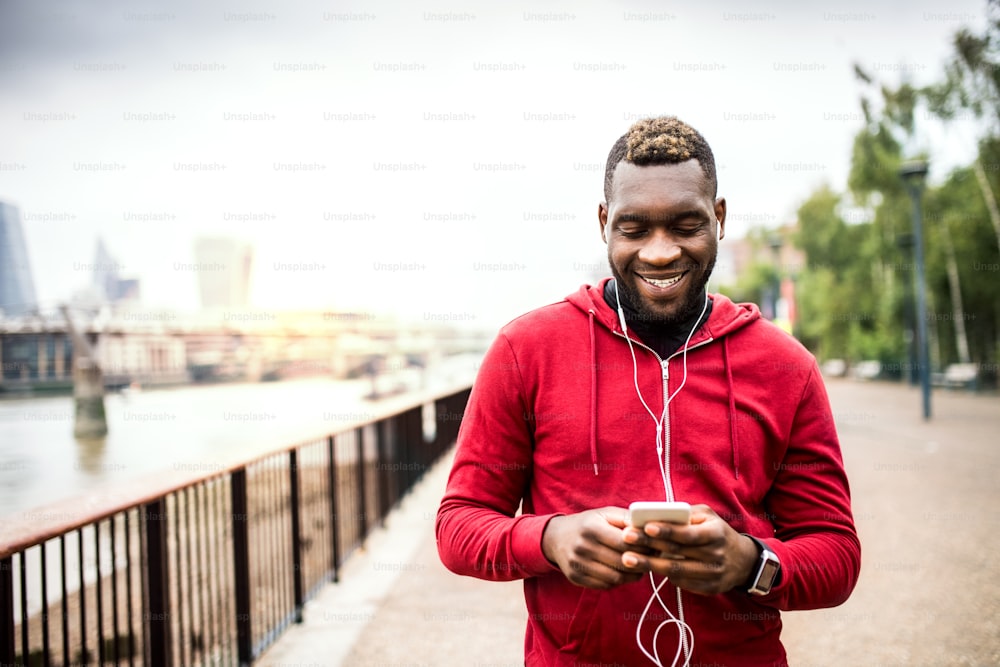 Joven corredor negro deportivo con auriculares, reloj inteligente y teléfono inteligente en el puente afuera en una ciudad, escuchando música.