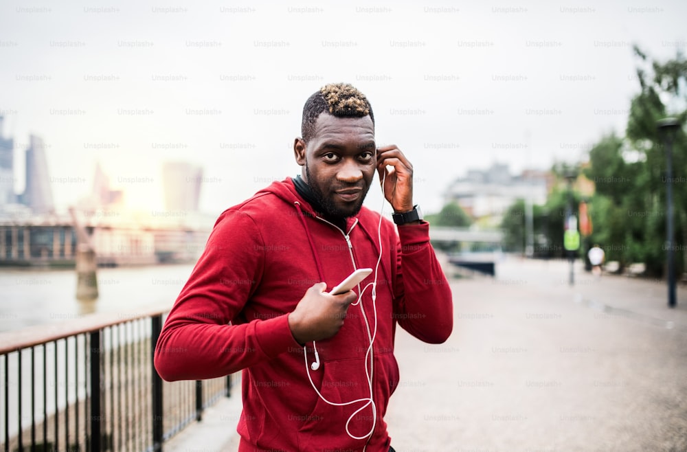 Junger sportlicher schwarzer Mann Läufer mit Kopfhörern, Smartwatch und Smartphone auf der Brücke draußen in einer Stadt, Musik hörend.