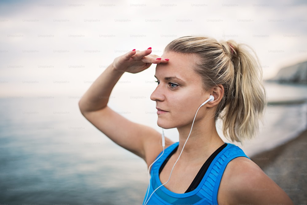 Young sporty woman runner with earphones standing on the beach outside, looking far away with her hand at the forehead.