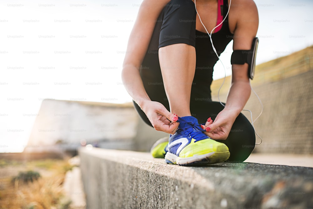 Unrecognizable young sporty woman runner with earphones and smartphone in an armband outside by the beach, tying shoelaces.