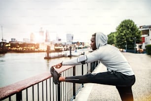 Young sporty black man runner with smartwatch, earphones and smartphone in an armband on the bridge in a city, stretching.