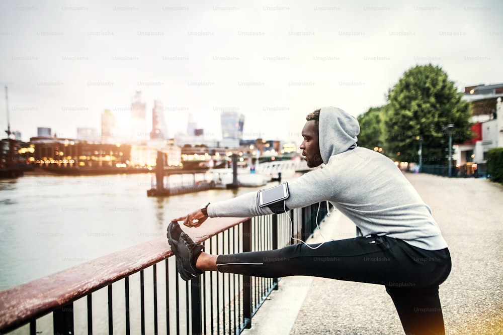 Jovem corredor negro esportivo com smartwatch, fones de ouvido e smartphone em uma braçadeira na ponte em uma cidade, se estendendo.