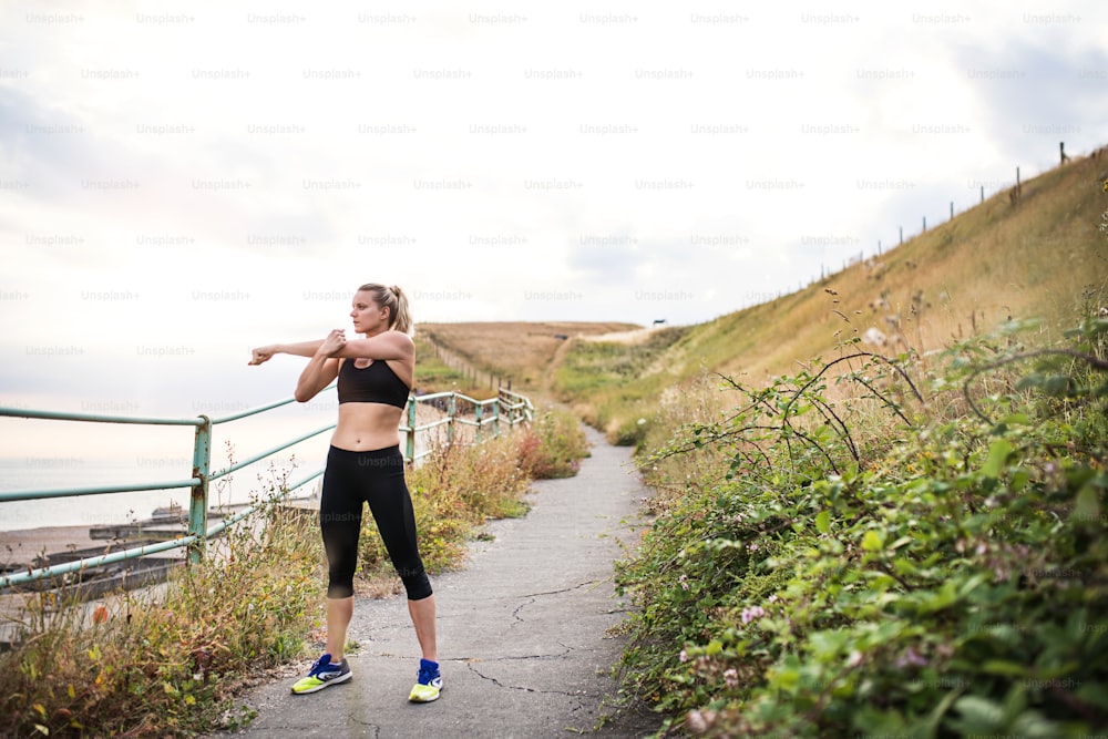 Young sporty woman runner in black activewear standing outside in nature, stretching. Copy space.