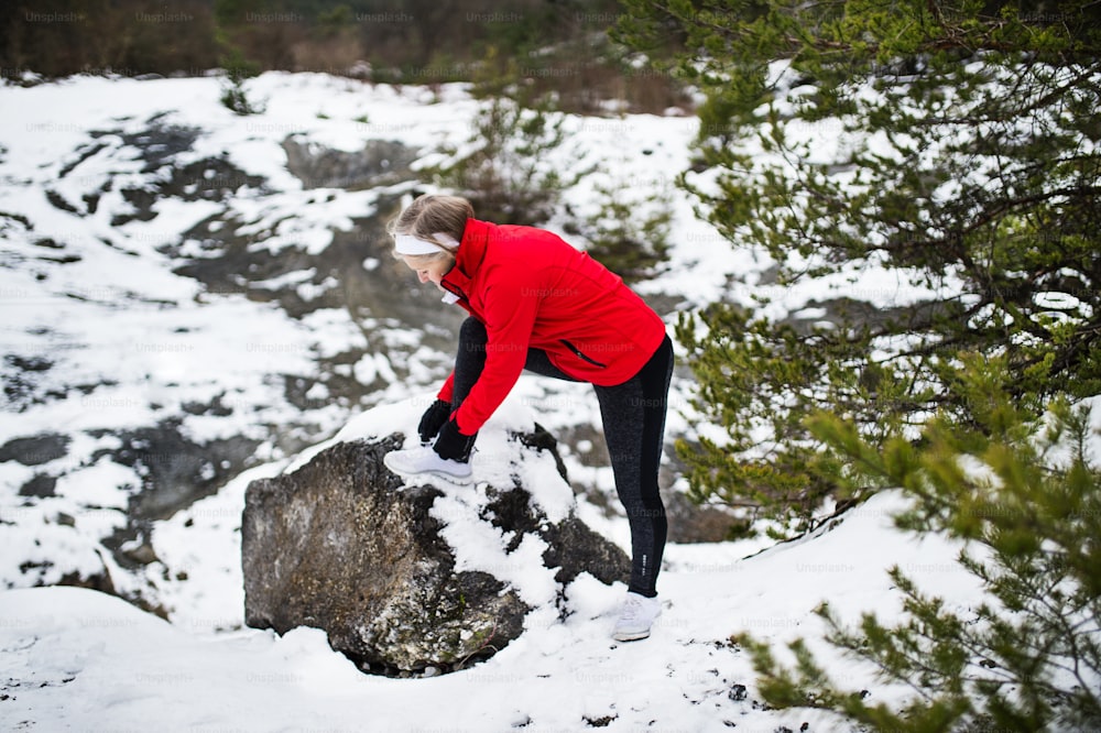 A senior woman runner tying shoelaces in winter nature, foot on rock.