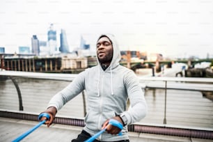Young active black sportsman exercising with elastic rubber bands on the bridge in London.