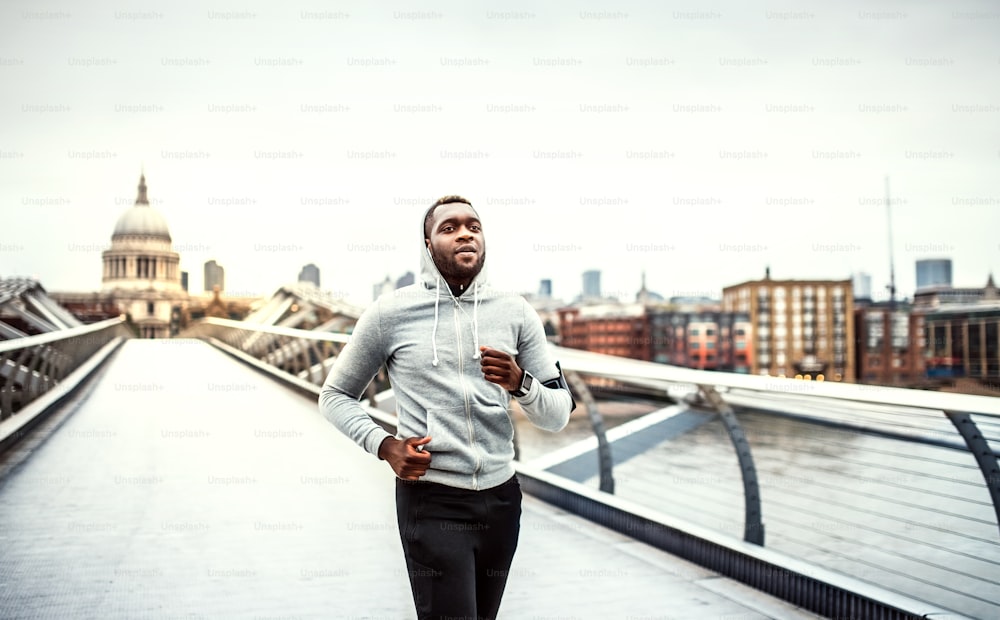 Junger sportlicher schwarzer Mann Läufer mit Smartwatch und Kopfhörern läuft auf der Brücke draußen in einer Stadt.