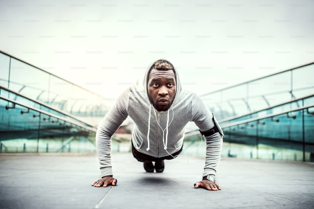 Young sporty black man runner with smartwatch and earphones doing push-ups on the bridge outside in a city.