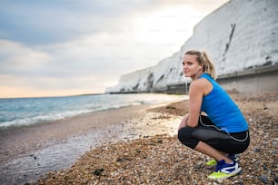 Young sporty woman runner with earphones resting outside on the beach in nature, listening to music. Copy space.