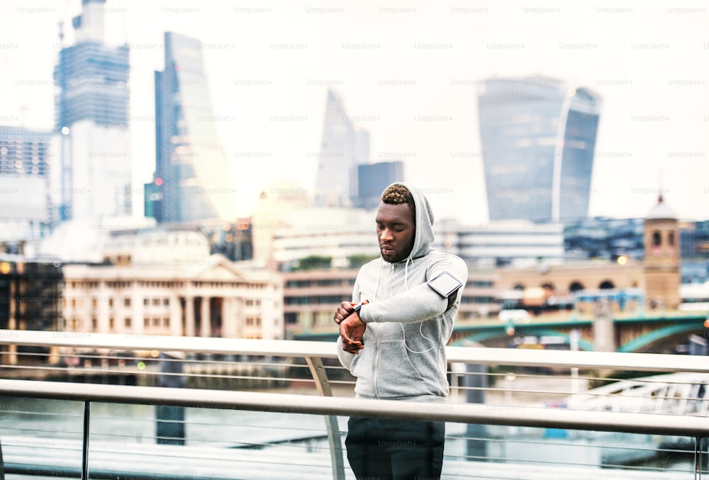 Young sporty black man runner with smartwatch, earphones and smartphone in an armband on the bridge in a city, resting.