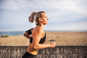 Jovem atleta mulher corredora com fones de ouvido correndo fora na praia na natureza, ouvindo música.