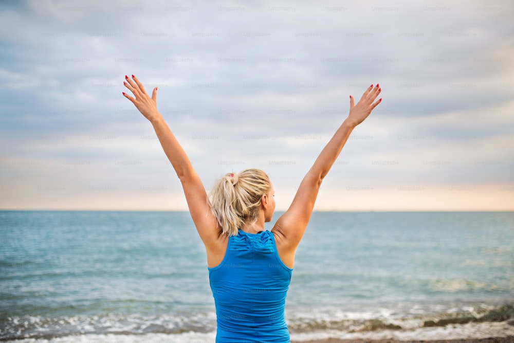 Jovem atleta mulher corredora em roupas esportivas azuis em pé fora na praia na natureza, descansando. Vista traseira.