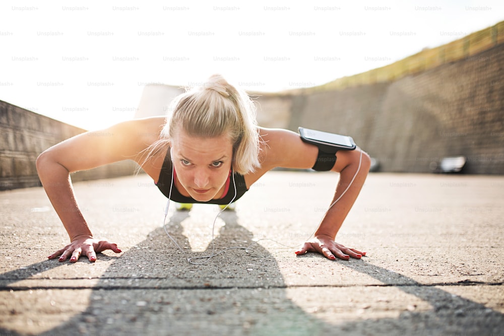 Young sporty woman runner with earphones and smartphone in armband doing push-ups outside in nature, listening to music.