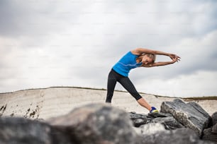 Young sporty woman runner with earphones standing outside on the rocks by beach in nature, listening to music and stretching. Copy space.