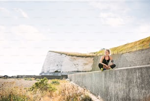 Young sporty woman runner with earphones and smartphone in armband sitting outside on the beach in nature, listening to music and resting.