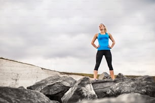 Young sporty woman runner with earphones standing outside on the rocks by beach in nature, listening to music and resting. Copy space.