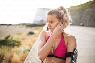 Joven corredora deportiva con auriculares y teléfono inteligente en el brazalete de pie afuera en la playa en la naturaleza, escuchando música y descansando.