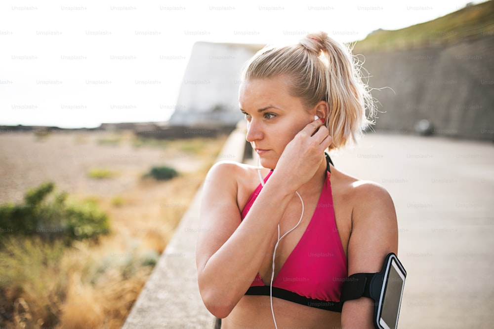 Young sporty woman runner with earphones and smartphone in armband standing outside on the beach in nature, listening to music and resting.