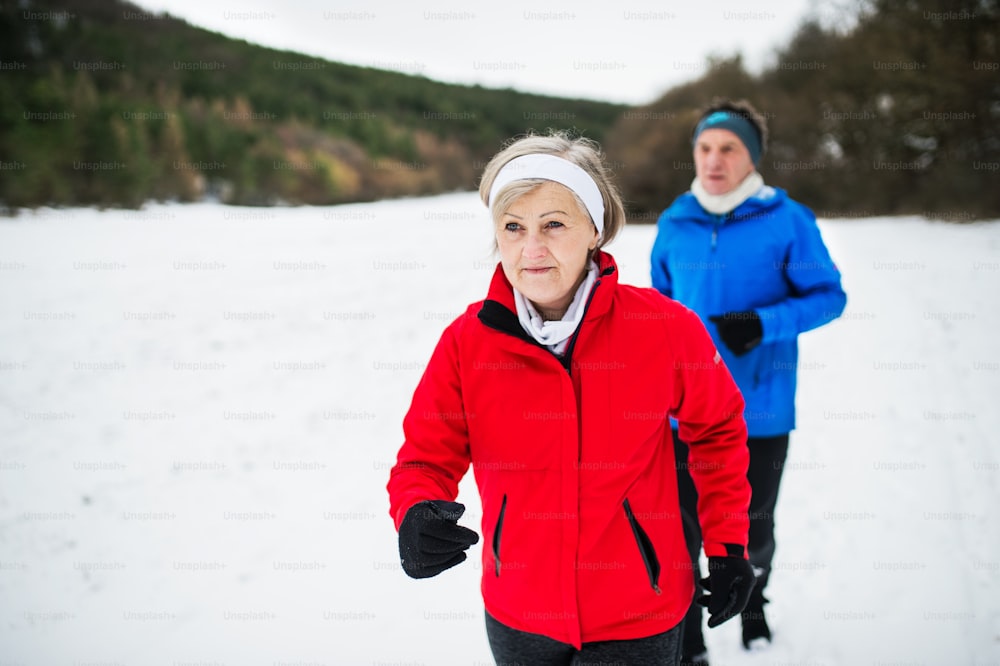 Una vista frontale di una coppia anziana felice che fa jogging nella natura invernale innevata.