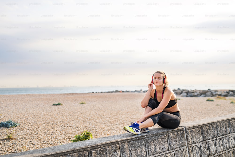 A young woman runner with black and red headphones resting outside by the sea, listening to music.
