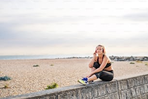 A young woman runner with black and red headphones resting outside by the sea, listening to music.