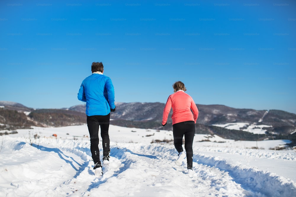 Vue arrière d’un couple de personnes âgées faisant du jogging dans la nature hivernale enneigée.