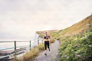 Junge sportliche Läuferin mit Kopfhörern, die draußen in der Natur am Meer entlang läuft und Musik hört.