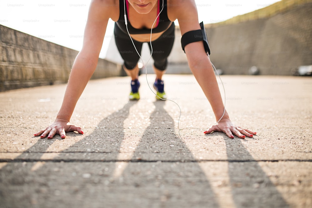 Unrecognizable young sporty woman runner with earphones and smartphone in armband doing push-ups outside in nature, listening to music.