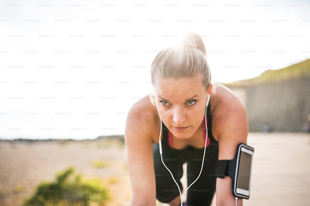 Young sporty woman runner with earphones and smartphone in armband standing outside on the beach in nature, listening to music and resting.