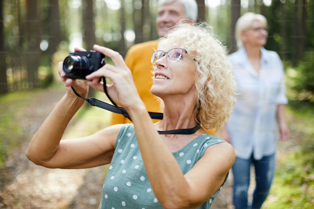 Smiling excited mature lady with curly hair wearing glasses looking up and photographing animal in forest while walking with friends