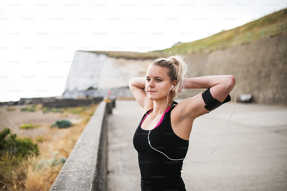 Young sporty woman runner with earphones and smartphone in armband standing outside on the beach in nature, listening to music and resting.