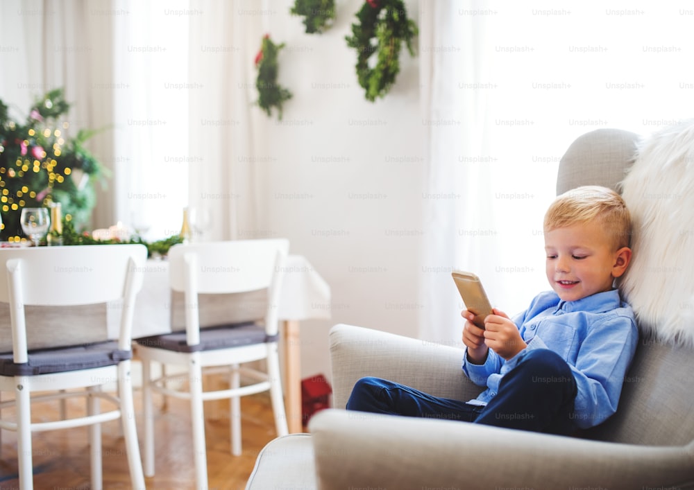 Un niño pequeño con un teléfono inteligente sentado en un sillón en casa en Navidad, jugando.
