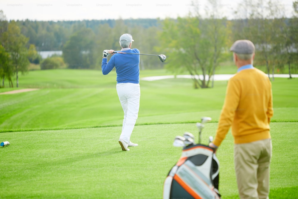 Aged man hit golf ball during leisure game while his buddy with bag for clubs standing near by