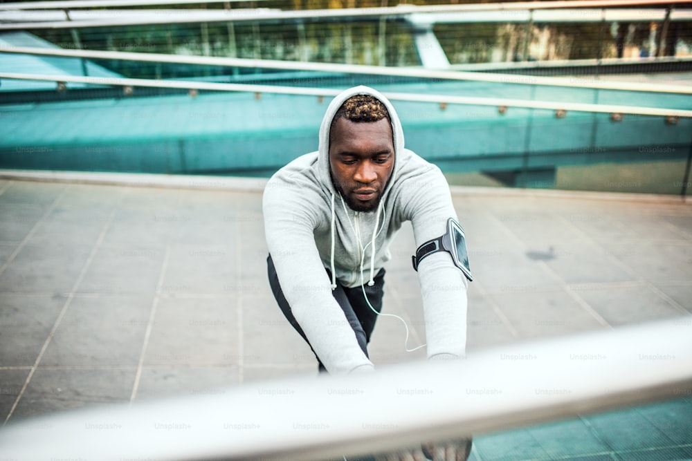 Young sporty black man runner with smartwatch, earphones and smartphone in an armband on the bridge in a city, stretching.