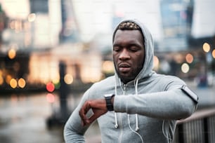 Young sporty black man runner with smartwatch, earphones and smartphone in an armband on the bridge in a London city, resting and measuring time.