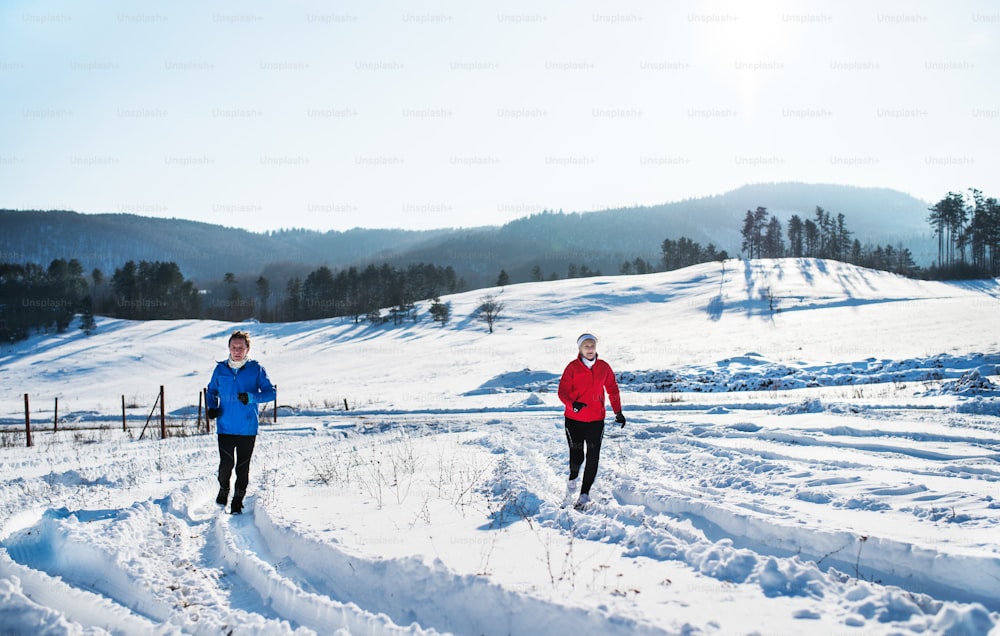 Une vue de face d’un heureux couple de personnes âgées faisant du jogging dans la nature hivernale enneigée.