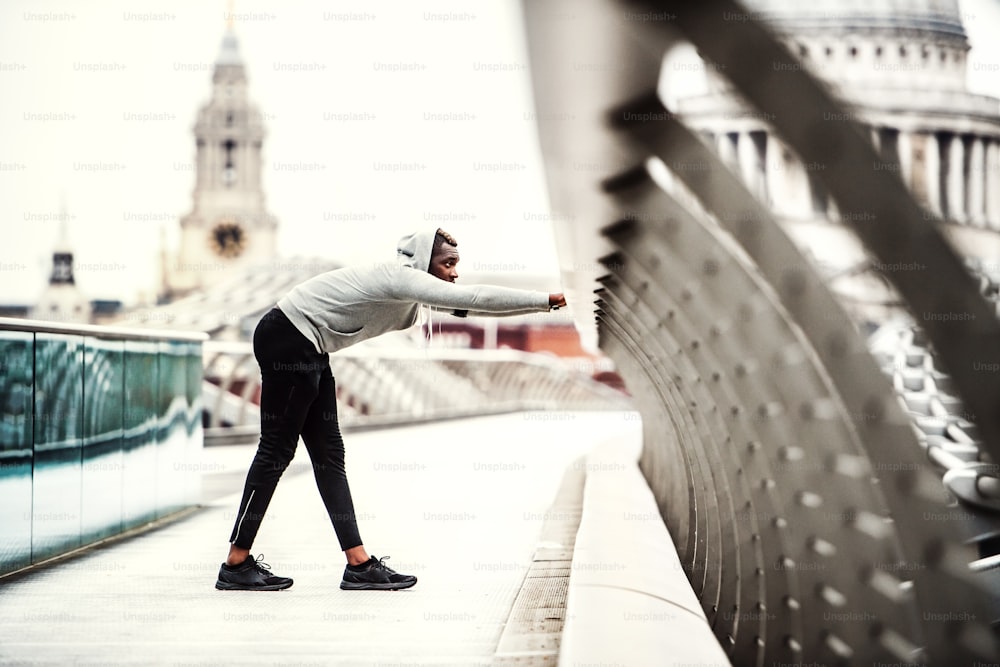 Young sporty black man runner on the bridge in a city, stretching.