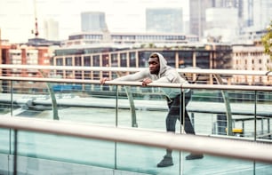 Jeune coureur noir sportif avec smartwatch, écouteurs et smartphone dans un brassard sur le pont d’une ville, en train de s’étirer.