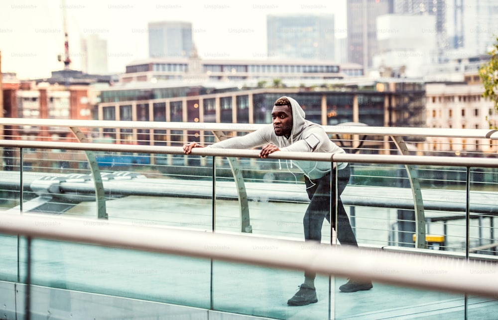 Junger sportlicher schwarzer Mann Läufer mit Smartwatch, Kopfhörern und Smartphone in einem Armband auf der Brücke in einer Stadt, streckend.