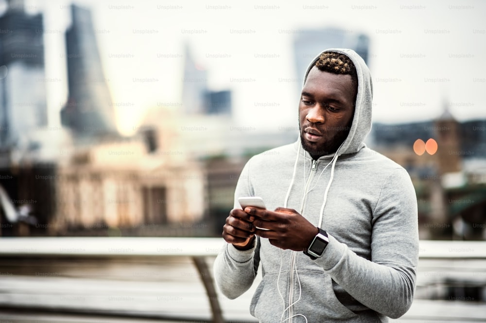 Young sporty black man runner with smartwatch, earphones and smart phone on the bridge in a city, resting. Copy space.