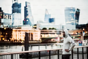 Young sporty black man runner with smartwatch, earphones and smartphone in an armband on the bridge in a city, resting. Copy space.