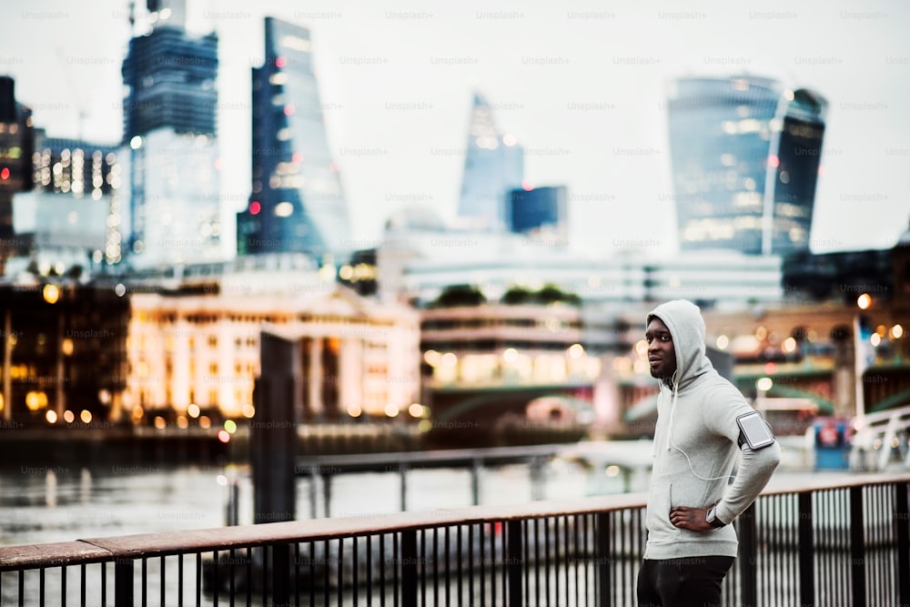 Young sporty black man runner with smartwatch, earphones and smartphone in an armband on the bridge in a city, resting. Copy space.