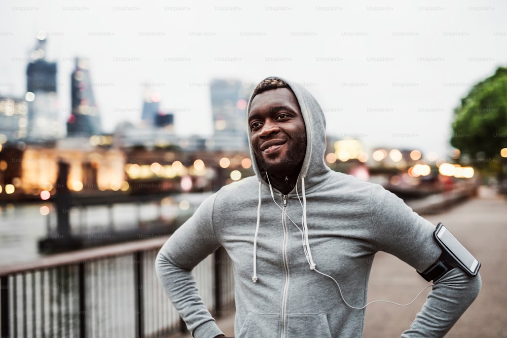 Jovem atleta negro corredor com smartwatch, fones de ouvido e smartphone em uma braçadeira na ponte em uma cidade, descansando.