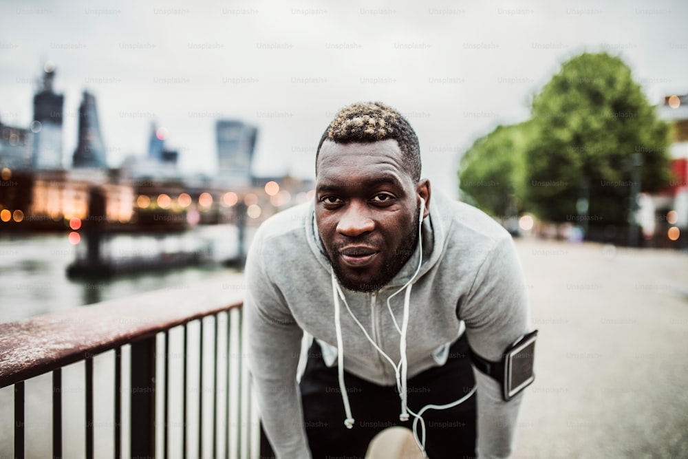 Young sporty black man runner with smartwatch, earphones and smartphone in an armband on the bridge in a city, resting. Copy space.