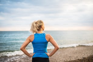 Giovane corridore sportivo donna in abbigliamento sportivo blu in piedi fuori sulla spiaggia nella natura, riposando. Veduta posteriore.