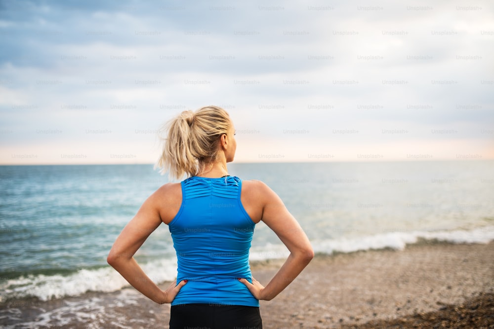 Junge sportliche Läuferin in blauer Sportbekleidung steht draußen am Strand in der Natur und ruht sich aus. Rückansicht.
