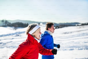 A side view of happy senior couple jogging in snowy winter nature.