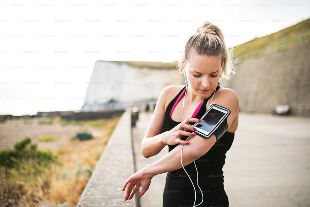 Jovem atleta mulher corredora com fones de ouvido em pé do lado de fora na praia na natureza, usando smartphone na braçadeira.
